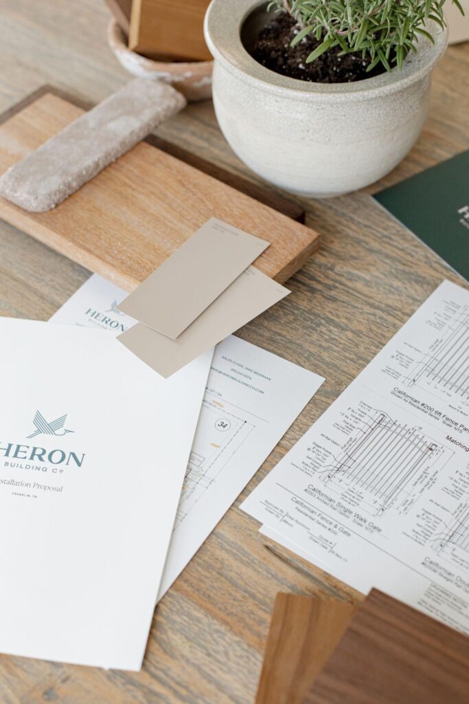 Fence design plans, material samples, and Heron Building Co. proposal documents laid out on a work table.