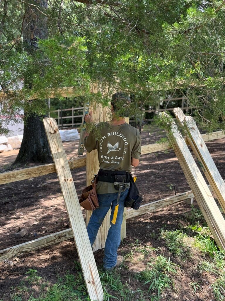 A Heron Building Co. installer levels cedar posts during a fence installation in a wooded Brentwood yard.