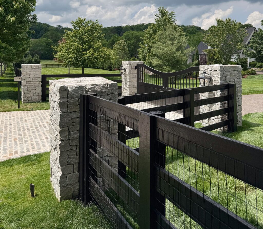 Black wood-and-wire fence with stone columns and an entry gate on a large estate.