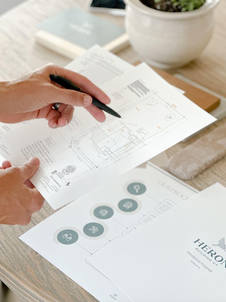 A person reviewing Heron Building Co. fence blueprints and project documents on a wooden table.
