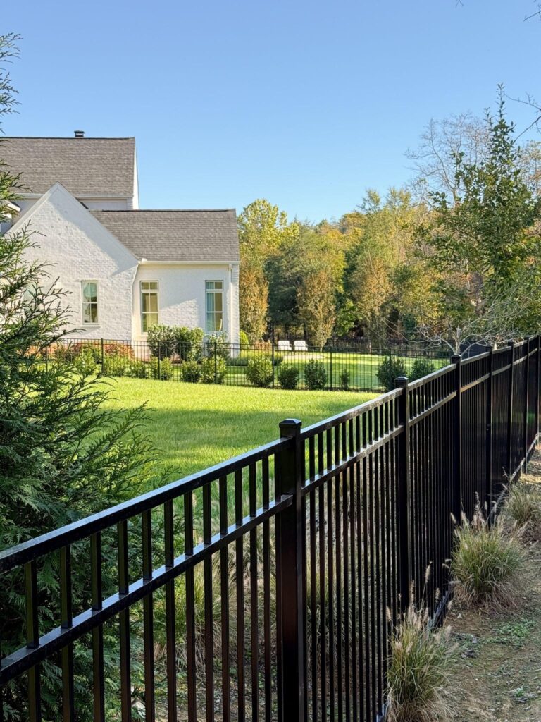 A sleek black aluminum fence bordering a Brentwood backyard with a white brick home and landscaped lawn.